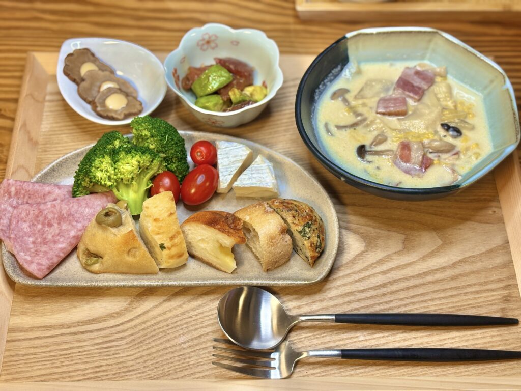 パンとチーズ、ビアソーセージ、ブロッコリー、トマトを盛り付けたおうちビストロ風のワンプレートと、ベーコンときのこの白菜クリーム煮、いぶりがっこチーズ、アボカドとマグロの山葵醤油和えの夕食セット。木のトレーの上に並べられた温かみのある家ごはん。
