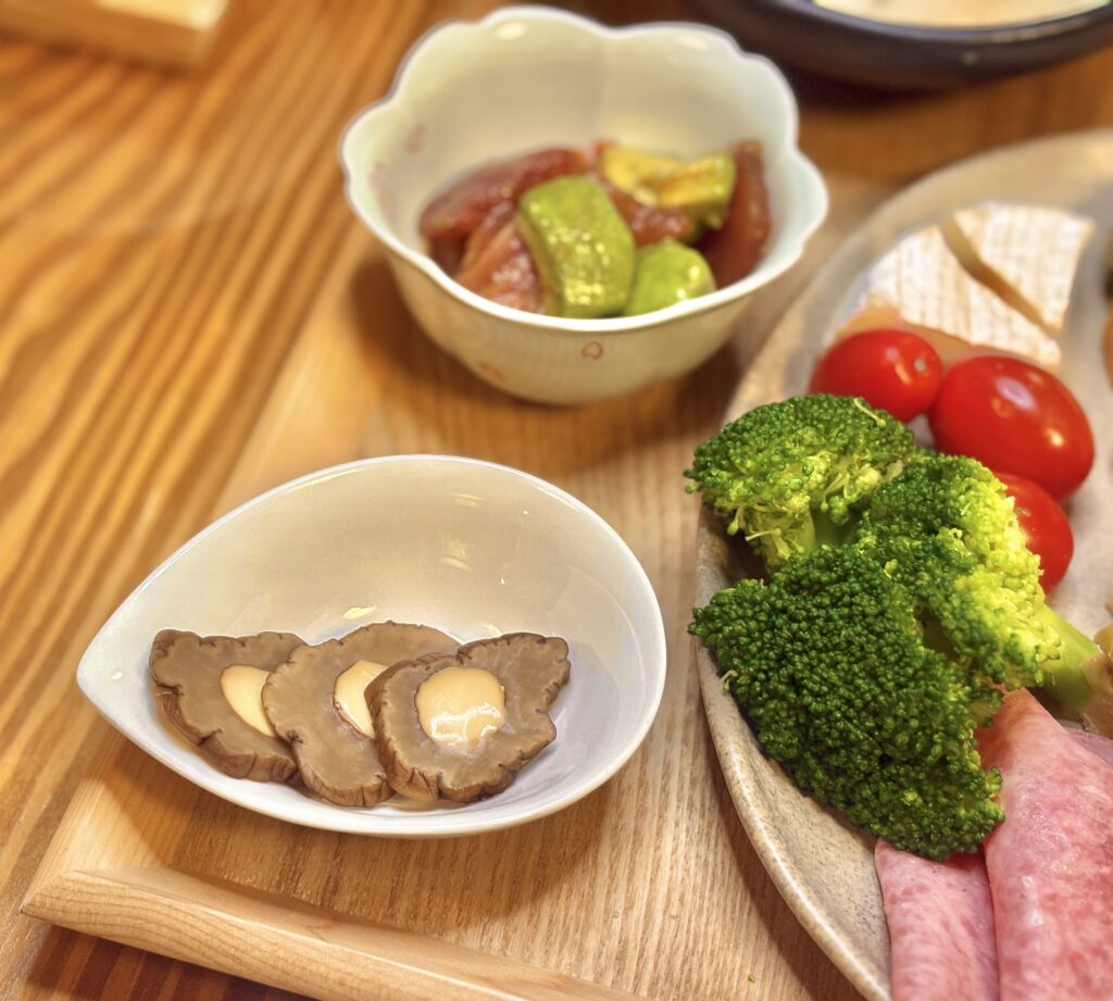 白い小鉢に盛られたいぶりがっこチーズと、奥にはアボカドとマグロの山葵醤油和えが入った器。隣にはブロッコリー、ミニトマト、カマンベールチーズ、ビアソーセージを添えたプレートの一部が見える。木のテーブルに並ぶおうちごはんの一皿。
