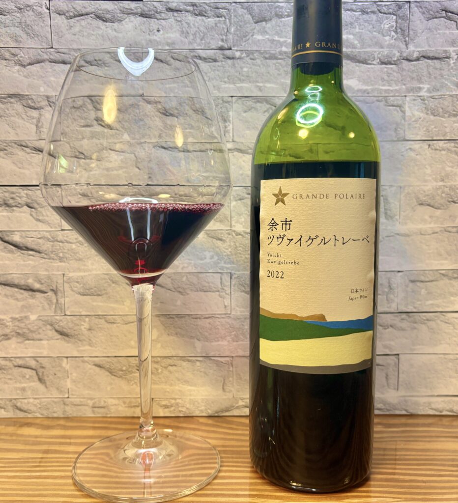 ワイングラスに注がれた赤ワインと、グランポレール「余市ツヴァイゲルトレーベ 2022」のボトル。木のテーブルの上に並べられ、背景には石壁風のグレーの壁。深いルビー色のワインが美しく、おうちディナーの雰囲気を演出している。