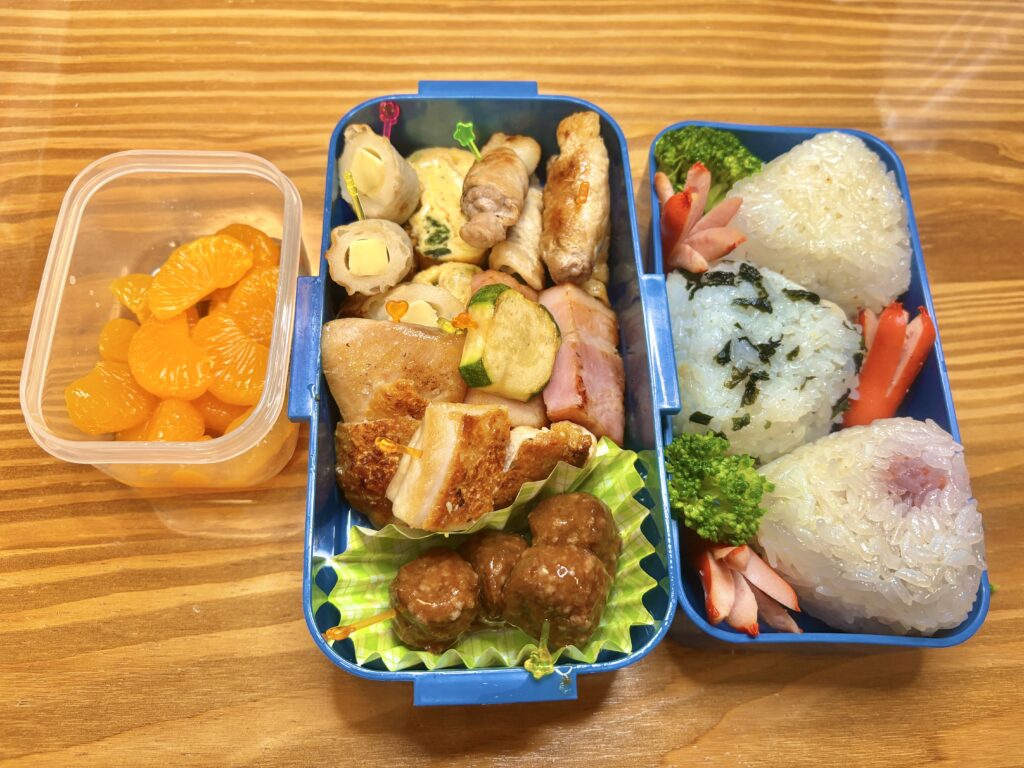 青いお弁当箱に、三角おにぎりとおかずが彩りよく詰められている。おかずには豚バラ巻き、ズッキーニのベーコン炒め、ミートボール、卵焼きなどが入り、別容器にはみかんのデザートが添えられている。