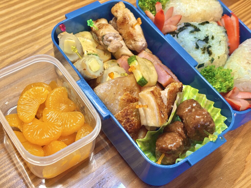 青いお弁当箱に詰められた手作り弁当。
左側には透明の容器に入ったみかんのデザート。
おかずには、照り焼き風ミートボール、鶏肉とズッキーニの串、
ベーコン、卵焼き、チーズちくわなどが彩りよく並び、
右側の段には梅おにぎり・わかめおにぎり・白おにぎりと、
ブロッコリーやタコさんウインナーが添えられている。
木目のテーブルの上で撮影された、あたたかい家庭の手作り弁当の写真。
