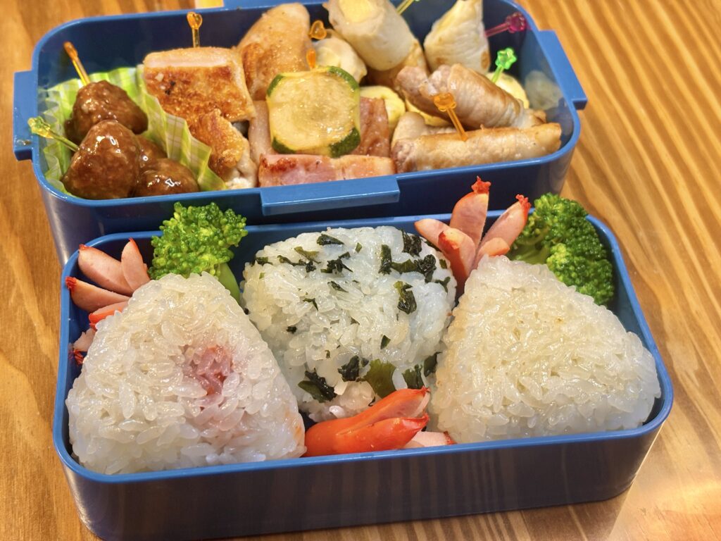 青いお弁当箱に詰められた手作り弁当。
下段には、梅おにぎり・わかめおにぎり・白おにぎりの3種類が並び、
彩りとしてタコさんウインナーとブロッコリーが添えられている。
上段には、照り焼き風のミートボール、鶏肉とズッキーニの串、
ベーコン炒め、卵焼き、豚肉の野菜巻きなどが彩りよく詰められており、
木目のテーブルの上で明るく撮影された、家庭的で温かみのあるお弁当の写真。