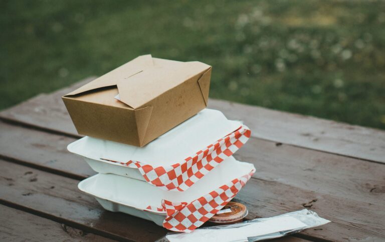 木製のピクニックテーブルの上に、テイクアウト用の容器が3段に重ねられて置かれている。 上には茶色のクラフト紙ボックス、下には白い容器が2つあり、 赤と白のチェック柄のワックスペーパーがはみ出している。 横にはフォークやスプーンが入ったビニール袋と、小さなソースカップも見える。 背景には芝生が広がっており、屋外での食事やピクニックを思わせる雰囲気の写真。