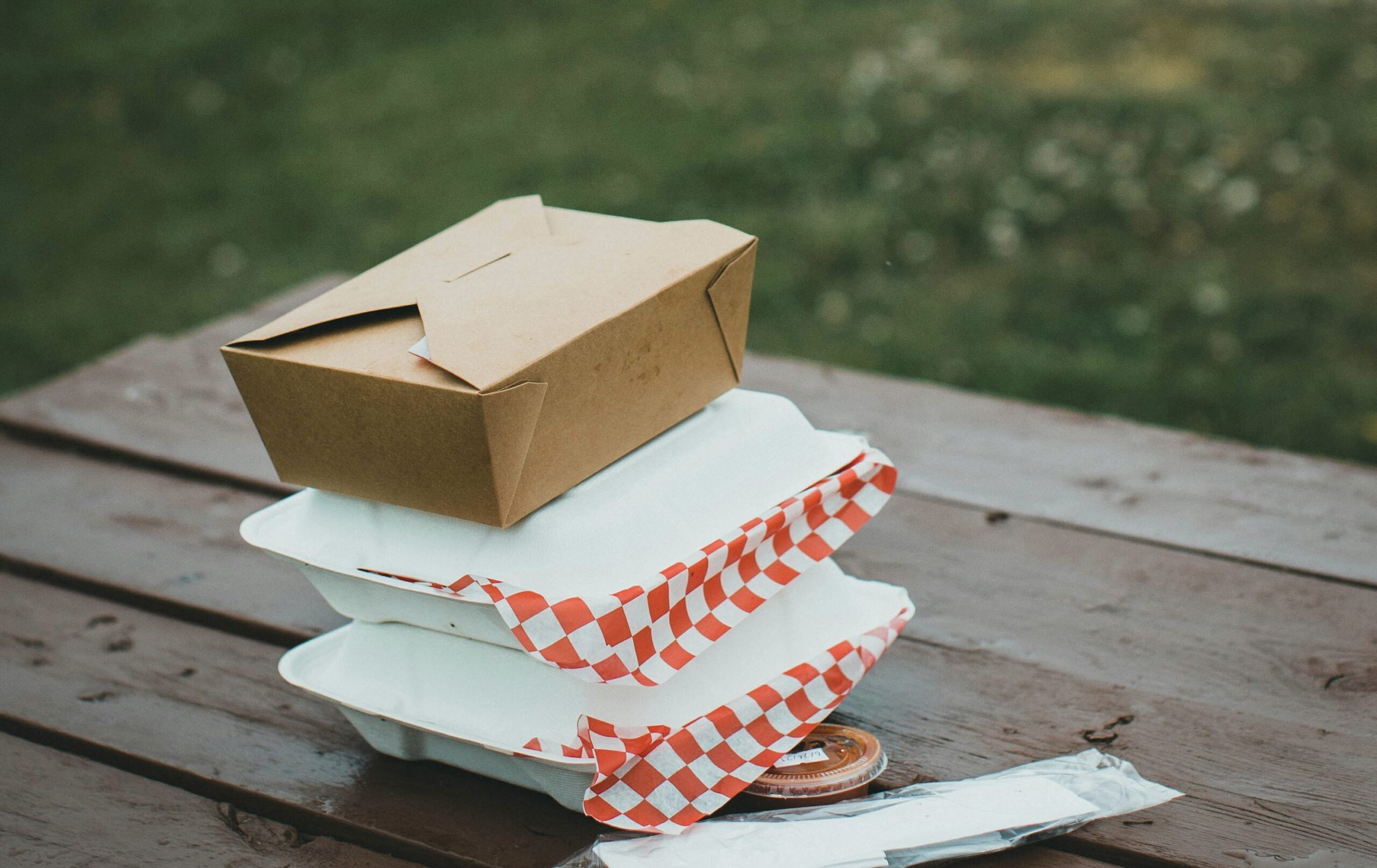 木製のピクニックテーブルの上に、テイクアウト用の容器が3段に重ねられて置かれている。 上には茶色のクラフト紙ボックス、下には白い容器が2つあり、 赤と白のチェック柄のワックスペーパーがはみ出している。 横にはフォークやスプーンが入ったビニール袋と、小さなソースカップも見える。 背景には芝生が広がっており、屋外での食事やピクニックを思わせる雰囲気の写真。