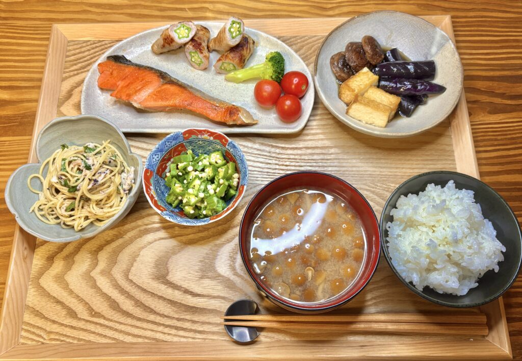 「木のトレイに並んだ和の晩ごはん。塩しゃけ、オクラの豚肉巻き、茄子と椎茸の揚げ浸し、オクラのおひたし、しらすと大葉のスパゲティ、なめこの味噌汁、ごはんを丁寧に盛りつけた家庭料理の食卓」