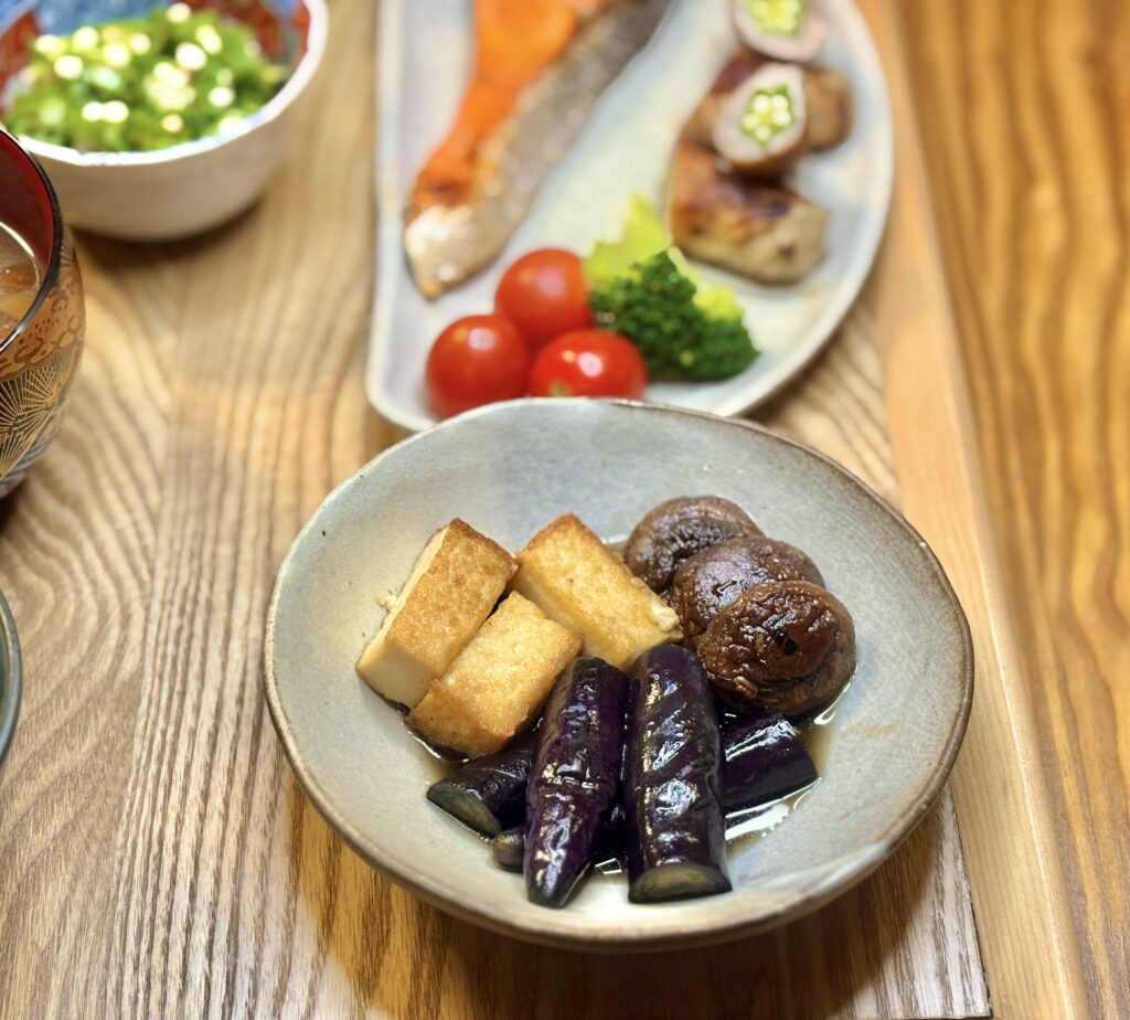 茄子の揚げ浸しに椎茸と厚揚げを添えた小鉢。奥には塩しゃけ、オクラの豚肉巻き、ブロッコリー、ミニトマトなどが並ぶ彩り豊かな和の晩ごはん