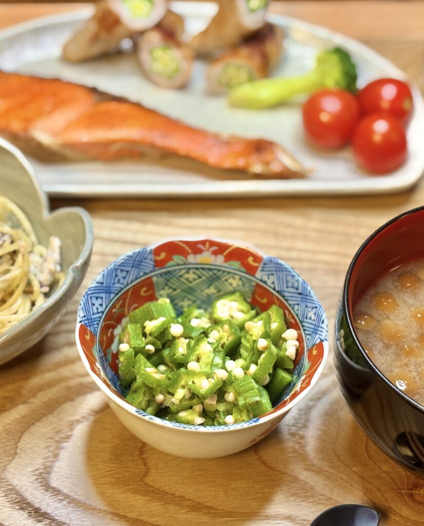 色鮮やかな和柄の小鉢に盛られた刻みオクラのおひたし。手前に味噌汁、奥には塩しゃけとオクラの豚肉巻き、ミニトマトなどが並ぶ和食の食卓