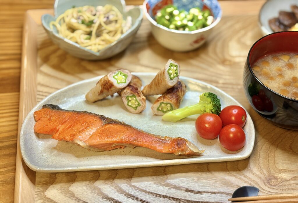 白い皿に盛りつけられた塩しゃけとオクラの豚肉巻き、ブロッコリーとミニトマトの彩り添え。奥にはオクラのおひたしやしらすのパスタ、手前にはなめこの味噌汁が並ぶ和の晩ごはん