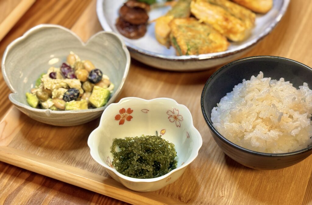 木のトレーに並べた家庭の和食ごはん。桜模様の器に盛られた海ぶどう、ハート型の器にアボカドと豆と鶏むね肉の胡麻ドレッシングあえ、黒い器の炊きたてごはんが並ぶ、温かみのある食卓。奥には黒皮カジキの青じそピカタが見える。
