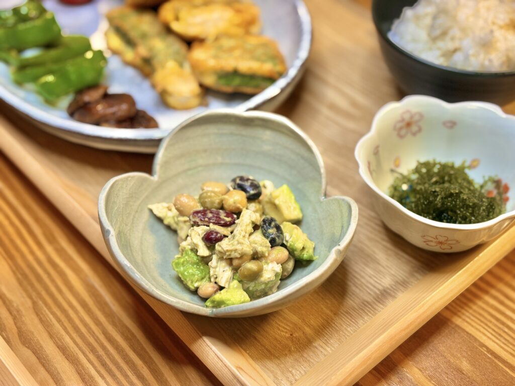 ハート型の器に盛られたアボカドと豆と鶏むね肉の胡麻ドレッシングあえ。奥には黒皮カジキの青じそピカタ、焼きピーマン、しいたけ、ミニトマト、ごはん、海ぶどうが並ぶ、木のトレーのおうちごはん。
