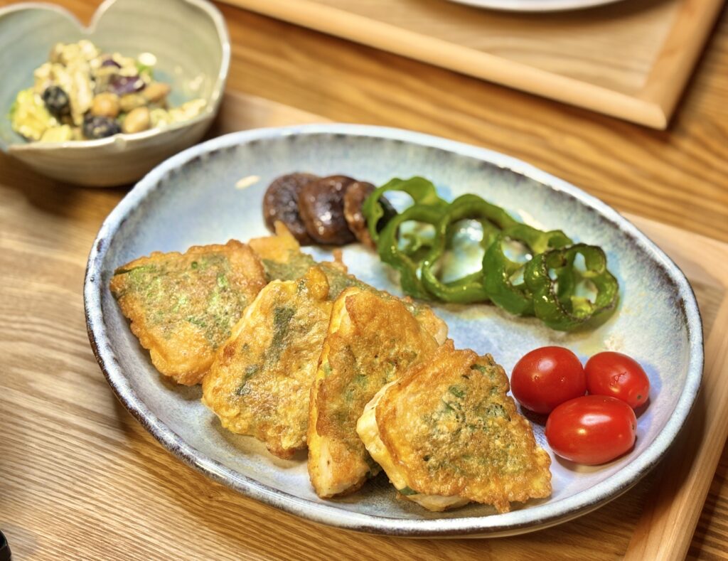 黒皮カジキの青じそピカタを中心に、焼きピーマン、焼きしいたけ、ミニトマトを添えたおうちごはんの一皿。奥にはアボカドと豆と鶏むね肉の胡麻ドレッシングあえが見える、木のテーブルの上の温かい食卓。
