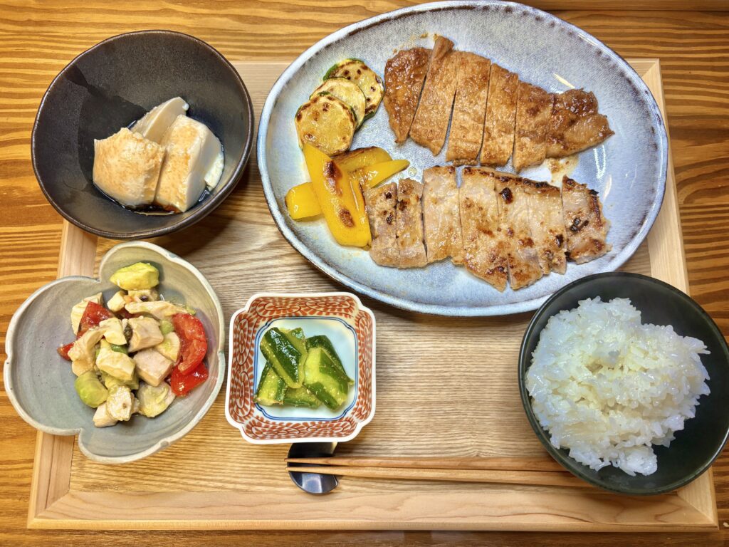 和豚もちぶたの生姜焼きと塩麹焼き、山芋とうふ、鶏胸肉とアボカドのサラダ、ごま油のたたききゅうりを並べた木製トレーの和食献立。