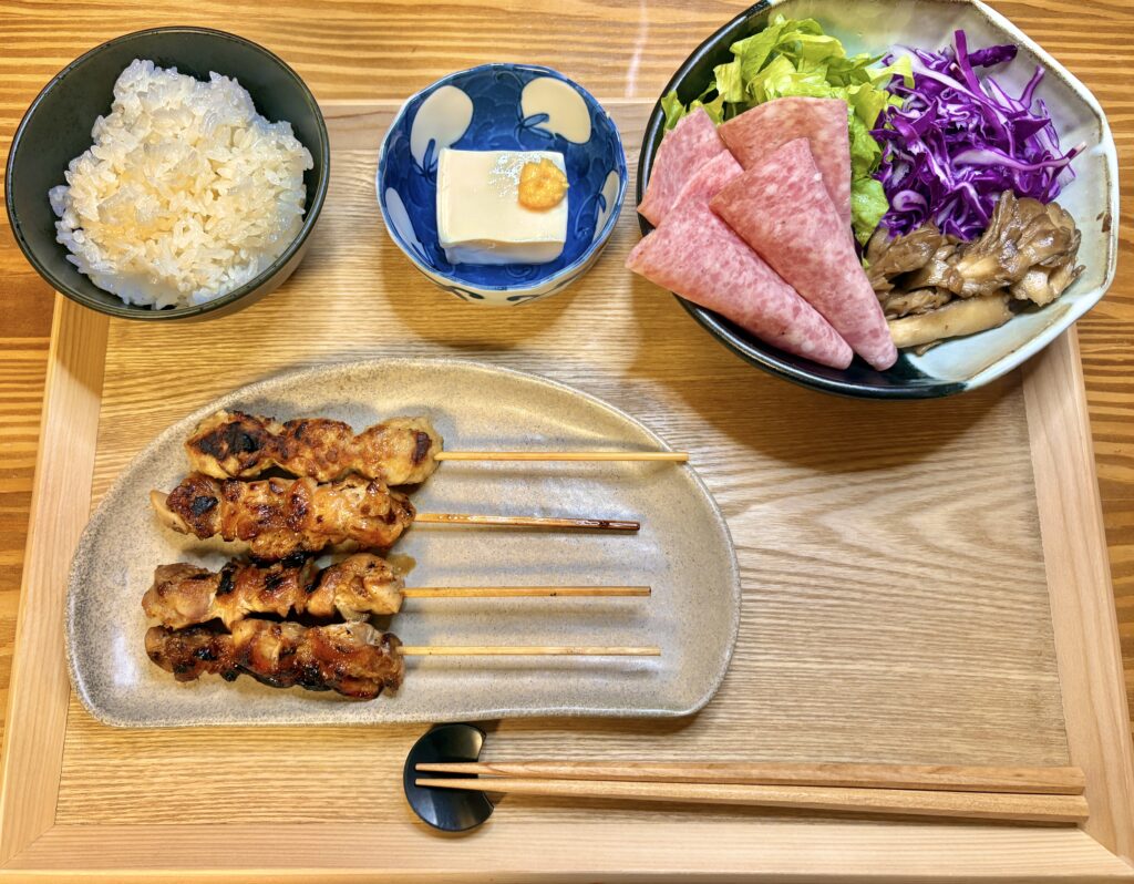 木のトレーに並べられた夕食セット。香ばしく焼かれた焼き鳥とつくね、ふっくら炊けた白ごはん、紫キャベツとビアソーセージの彩りサラダ、冷奴が並ぶ家庭の食卓。