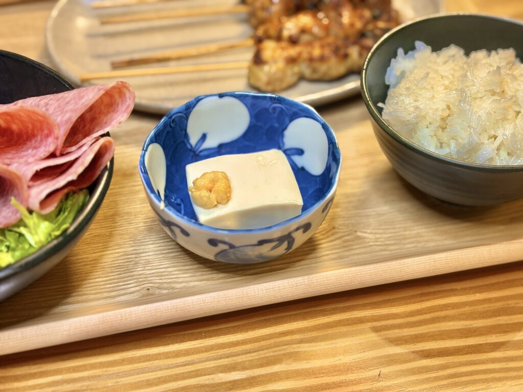 青い模様の小鉢に盛られた冷奴。上におろし生姜がのり、隣には白ごはんとビアソーセージのサラダが並ぶ、家庭的で温かみのある和食の食卓。