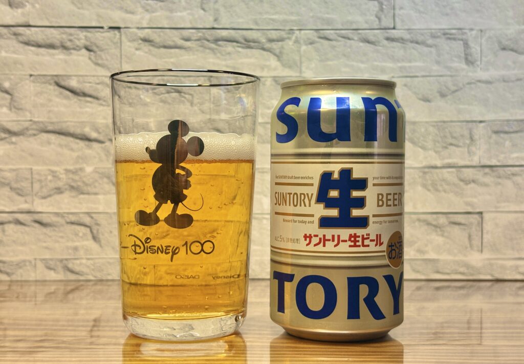 ミッキーのシルエットが描かれたグラスに注がれた黄金色のビールと、横に並ぶサントリー生ビールの缶。明るい木目のテーブルと白いレンガ調の背景で、シンプルにおしゃれな乾杯シーンが写っている。