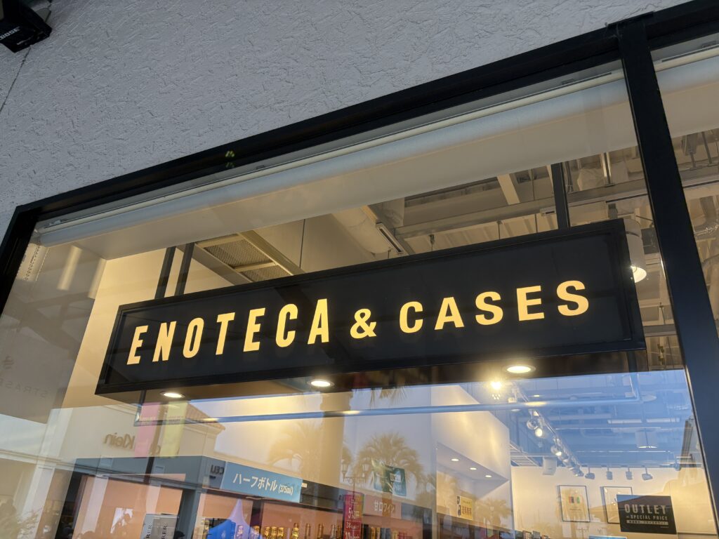 エノテカ&ケイセス(ENOTECA & CASES)の店舗外観。黒い看板に黄色い文字で店名が大きく表示され、その下にはガラス張りの明るい店内が見えている。照明がきらめき、ワインショップらしい洗練された雰囲気が伝わる写真。ガラスには周囲のアウトレットモールの景色や人影が反射している。