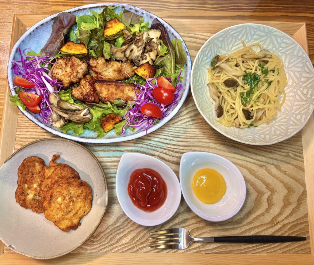 彩りたっぷりのグリルチキンサラダ、しめじのペペロンチーノ、手作りチキンナゲットが木のトレイに並んだ晩ごはんの写真。