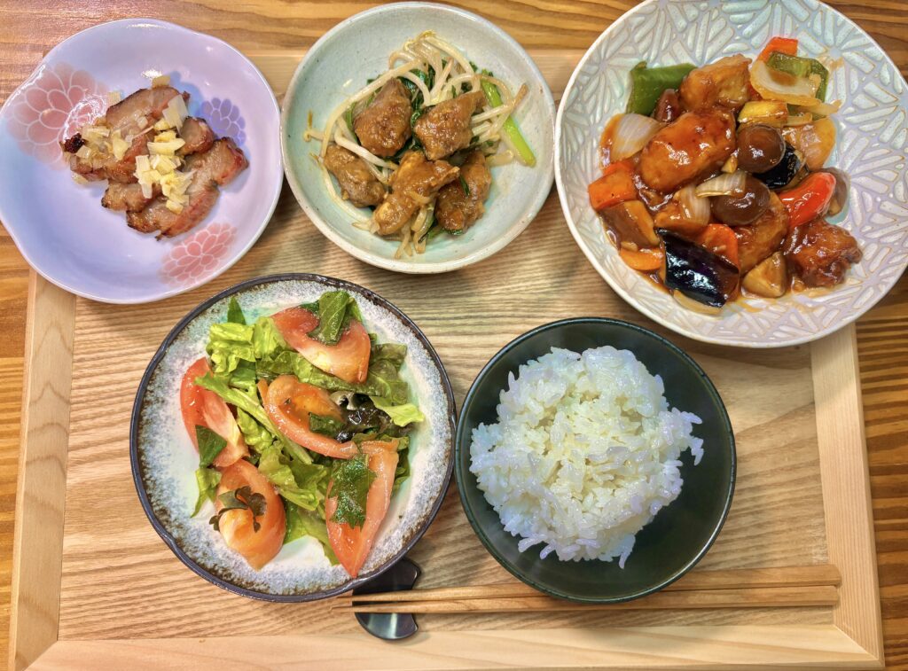 木製トレイに、ご飯、トマトと葉野菜のサラダ、酢豚風のおかず、野菜炒め、小皿の焼き豚を並べた和洋中ミックスの食卓。色鮮やかな惣菜がバランスよく盛り付けられている。