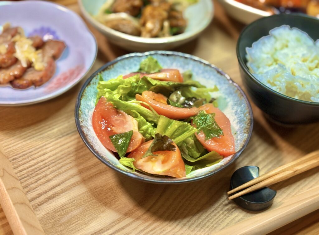 青みがかった器に、レタスとトマトを盛り付けたフレッシュなサラダが入っている。横には黒い器の白ご飯、後ろには焼き豚や炒めものが並び、木のトレイの上に和洋中がそろった食卓が広がっている。