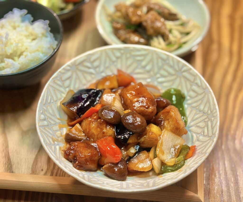 白い模様入りの皿に、なす・玉ねぎ・ピーマン・パプリカと一緒に豚肉を甘酢あんで炒めた中華風おかずが盛られている。具材にツヤがあり、色鮮やかな一皿。