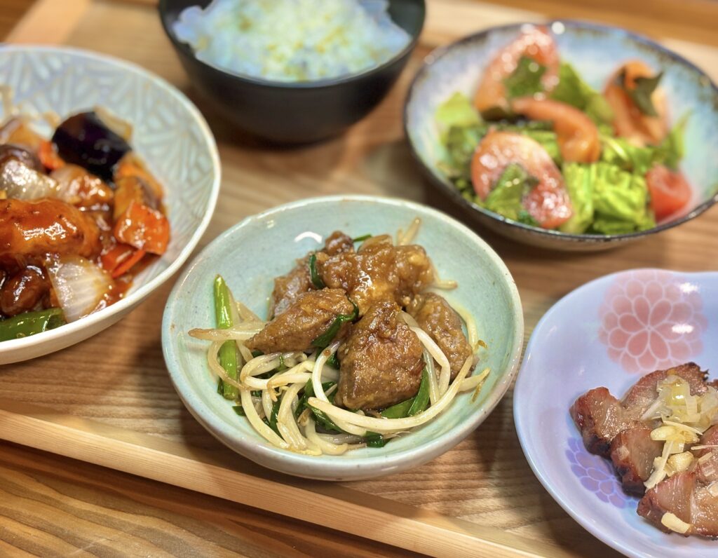 薄い緑色の器に、豚肉とニラともやしを炒めた中華風のおかずが盛られている。周りにはサラダや酢豚、小皿の焼き豚が並び、木のトレイの上に彩りよく配置されている。