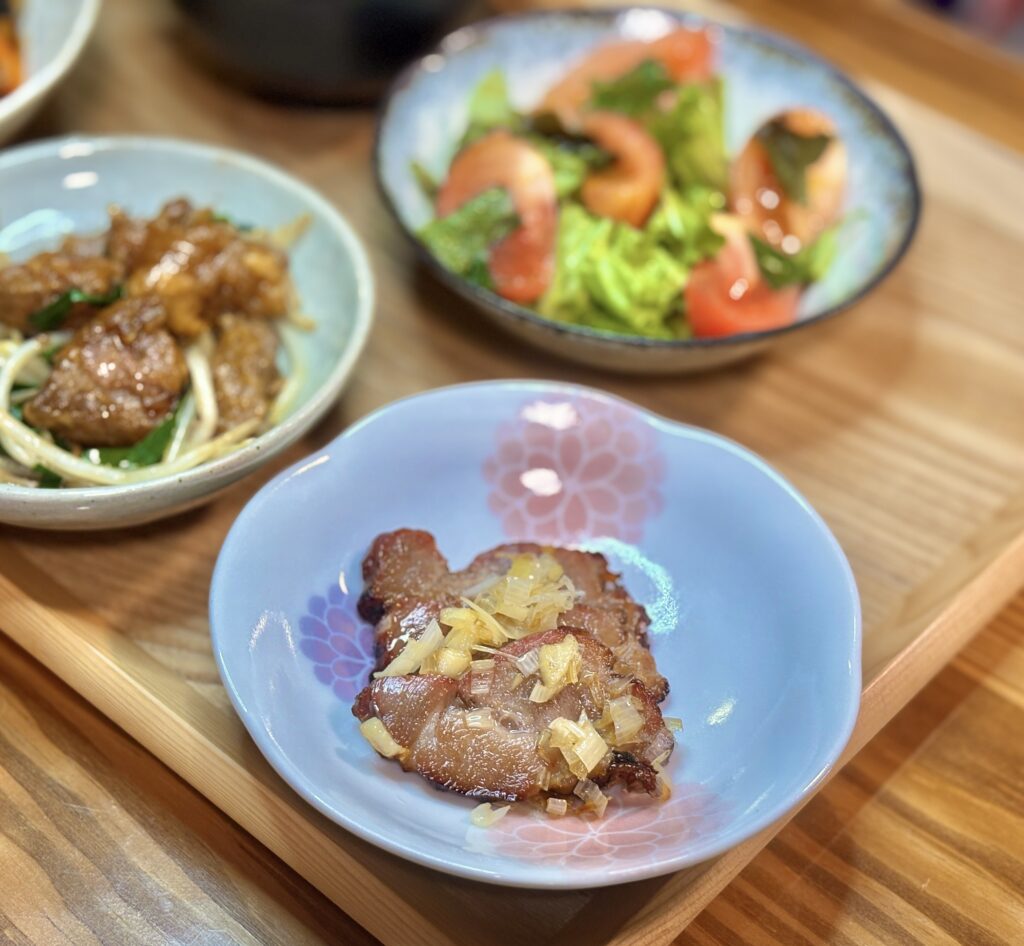 花柄の小皿に、薄切りの焼き豚の上から刻んだねぎの香味だれをかけた一品が盛られている。背景にはサラダや炒めものがぼんやりと写り、木のトレイの上で食卓の一部が切り取られている。