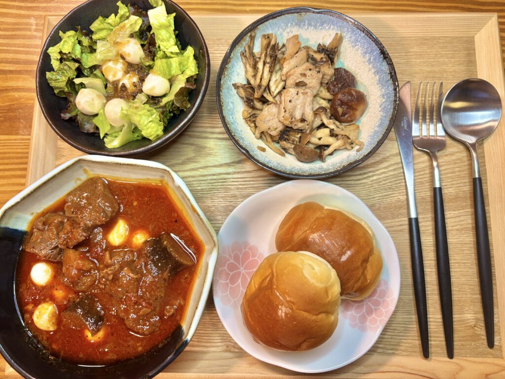 木目のトレイの上に、牛肉のトマト煮込み、キハダマグロときのこのアヒージョ、モッツァレラサラダ、丸パンが並んだ洋食ディナーセットが美しく配置されている。左上にサラダ、右上にアヒージョ、左下に煮込み、右下にパンが置かれ、横には黒い持ち手のフォークとスプーンが揃えて並べられている。