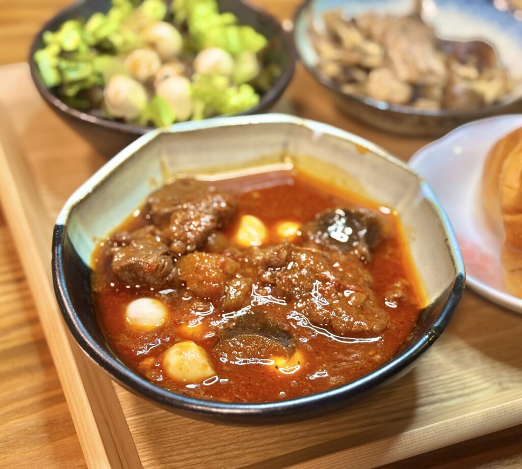 深みのある赤いソースに包まれた牛肉の塊とモッツァレラチーズが、艶やかな表面で光を反射している。器の縁に焦点が合い、背景にはサラダやパンがぼんやりと映り込む。煮込みの濃厚さと温かさが伝わる質感が際立つ一枚。