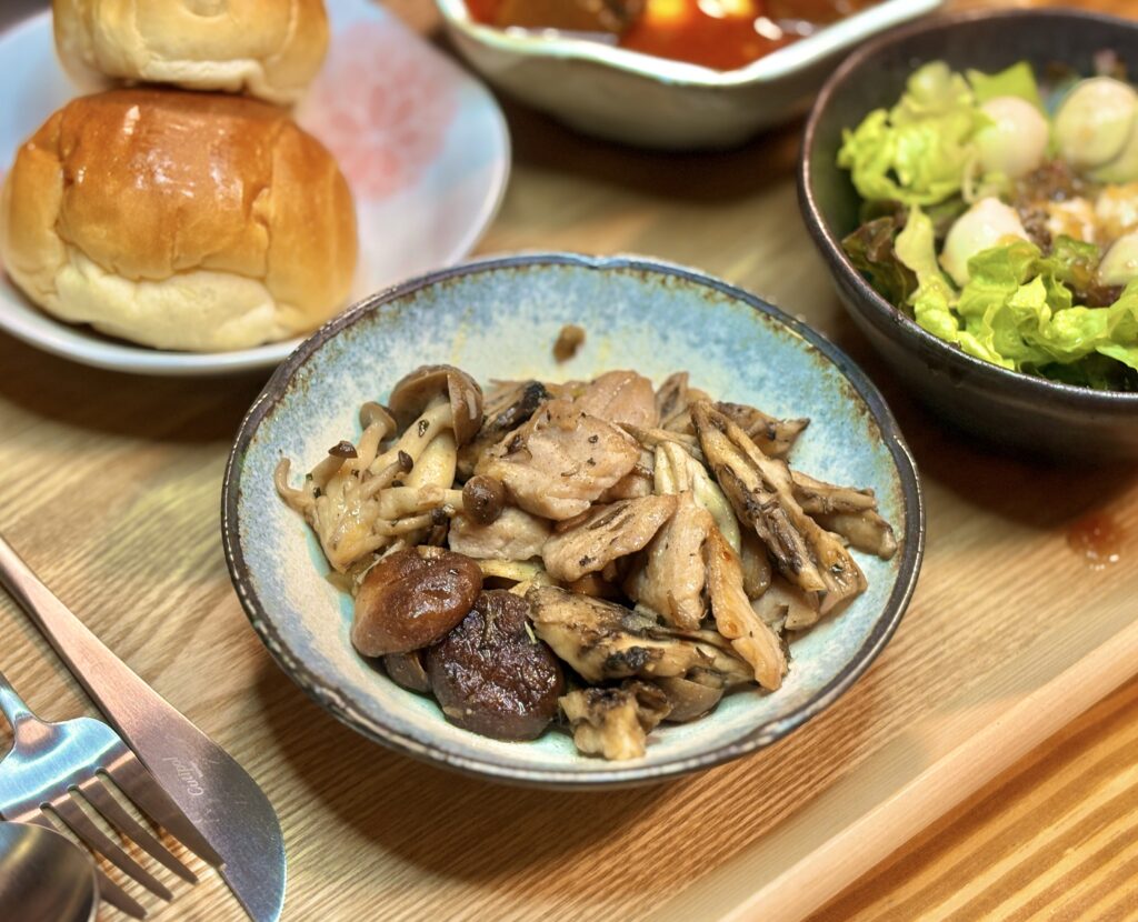 青みがかった和陶器の皿に、しめじやまいたけ、しいたけのきのこと、しっとり火が入ったキハダマグロが盛り付けられている。木目のトレイの上で、きのこの香りが立つような温かみのある質感が伝わり、手前のカトラリーが少し映り込んでいる。