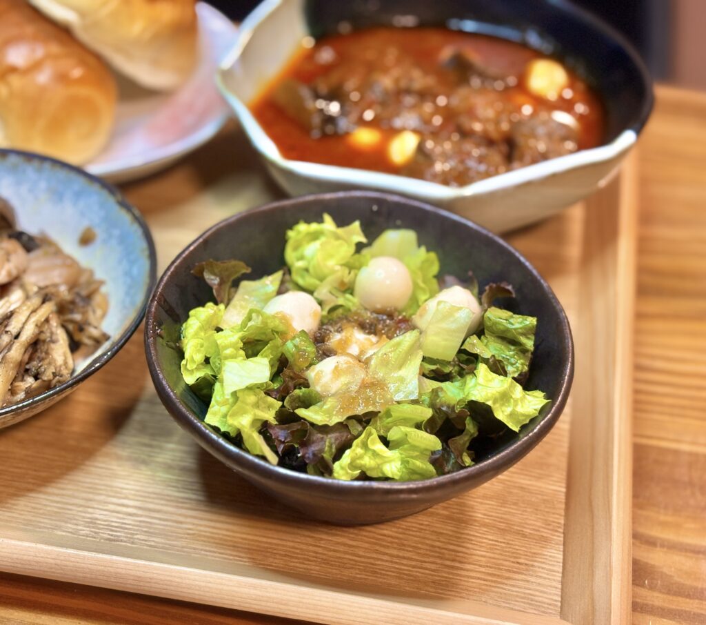 黒い器に、明るいグリーンのレタスと白いモッツァレラチーズの丸い形が映えるフレッシュなサラダ。中央にはドレッシングがかかり、光が葉に反射してみずみずしさを強調している。背景にはトマト煮込みの赤がぼんやりと写り、食卓の統一感が出ている。