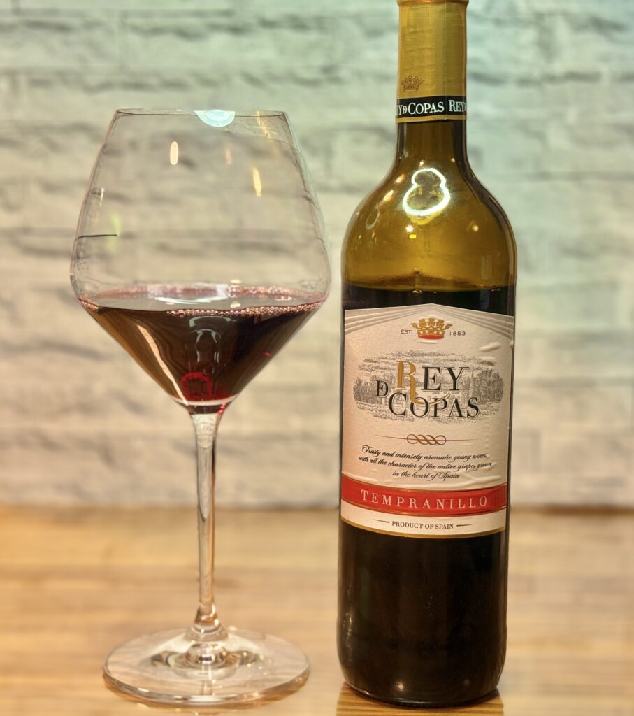 REY DE COPAS の赤ワインボトルと、グラスに注がれたルビー色のワインが並ぶ一枚。白いレンガ調の背景に、ワインの色味が美しく際立ち、グラスの曲線に照明の反射がやわらかく映っている。落ち着いた雰囲気で、食卓の締めくくりを感じさせる構図になっている。