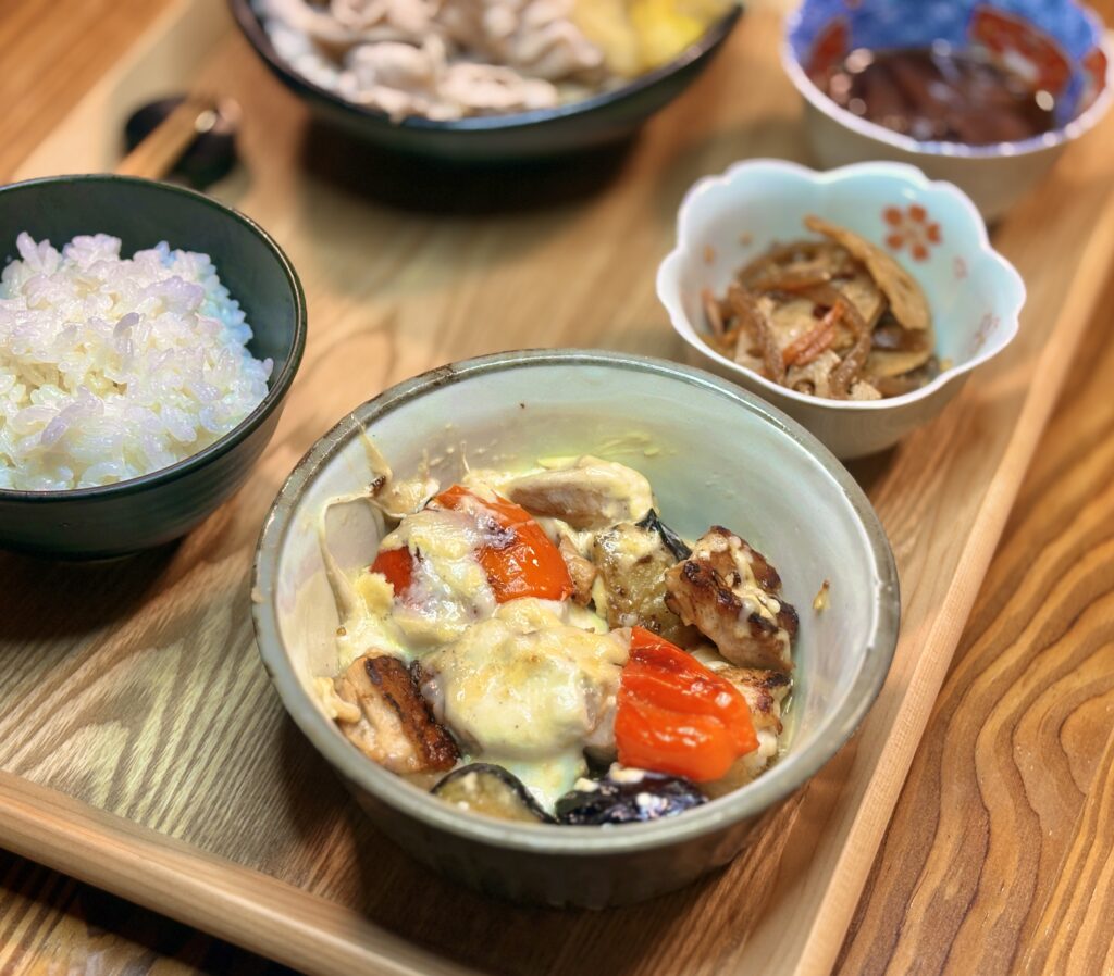 副菜の「茄子とパプリカのガーリック炒め」のクローズアップ写真です。ジューシーな揚げ茄子と鮮やかな赤パプリカに、とろりとしたソースやチーズが絡み合い、ガーリックの香ばしい風味が漂ってきそうな、食欲をそそる一皿です。
