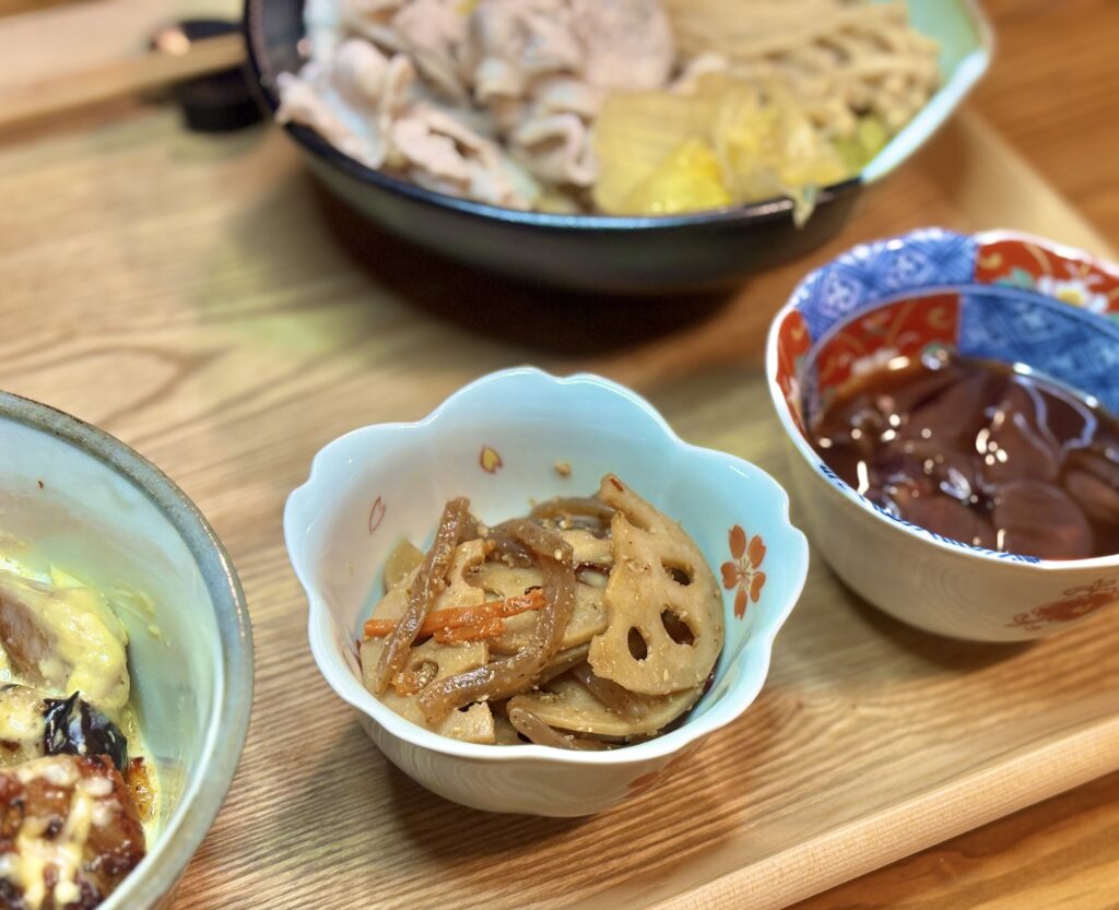 副菜の「蓮根と人参のきんぴら」のクローズアップ写真です。白い花びらの形をした可愛らしい小鉢に、胡麻が振られた蓮根と人参が丁寧に盛り付けられています。甘辛い味付けとシャキシャキとした食感が想像でき、箸休めにぴったりの定番和惣菜です。
