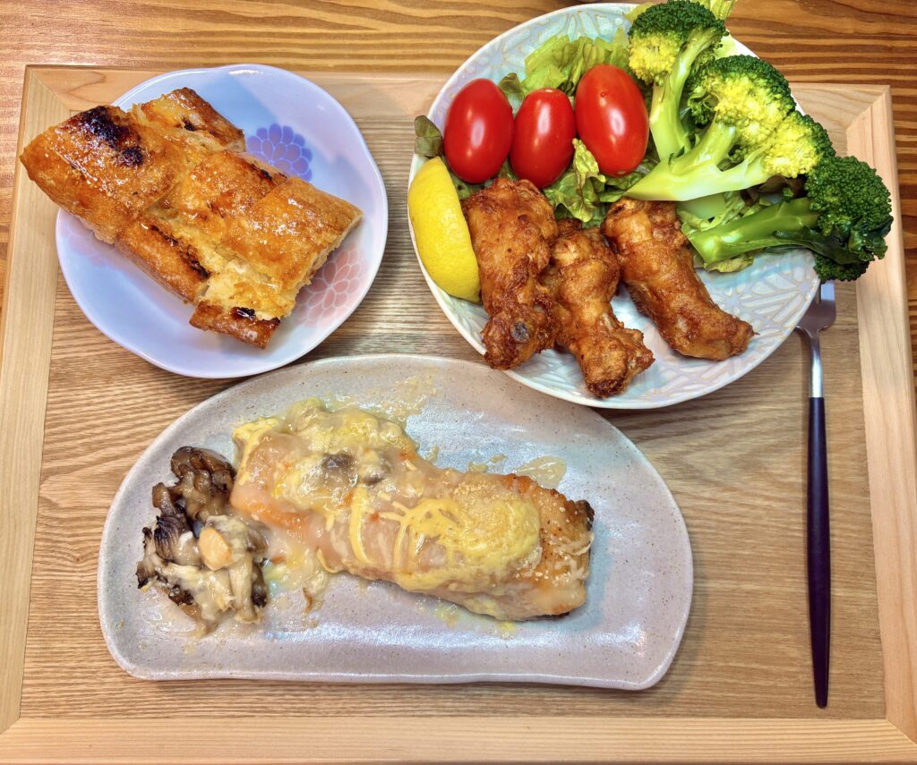 フライドチキン、ブロッコリーとミニトマトのサラダ、サーモンと舞茸のホワイトソースチーズ焼き、メイプルバターバゲットが木のトレイに並んだ夕食プレート。
