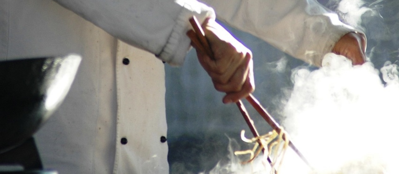 白いコックコートを着た人が、大きな箸で麺を炒めている様子。立ちのぼる湯気の中で、手元の動きが強調された臨場感あるカット。