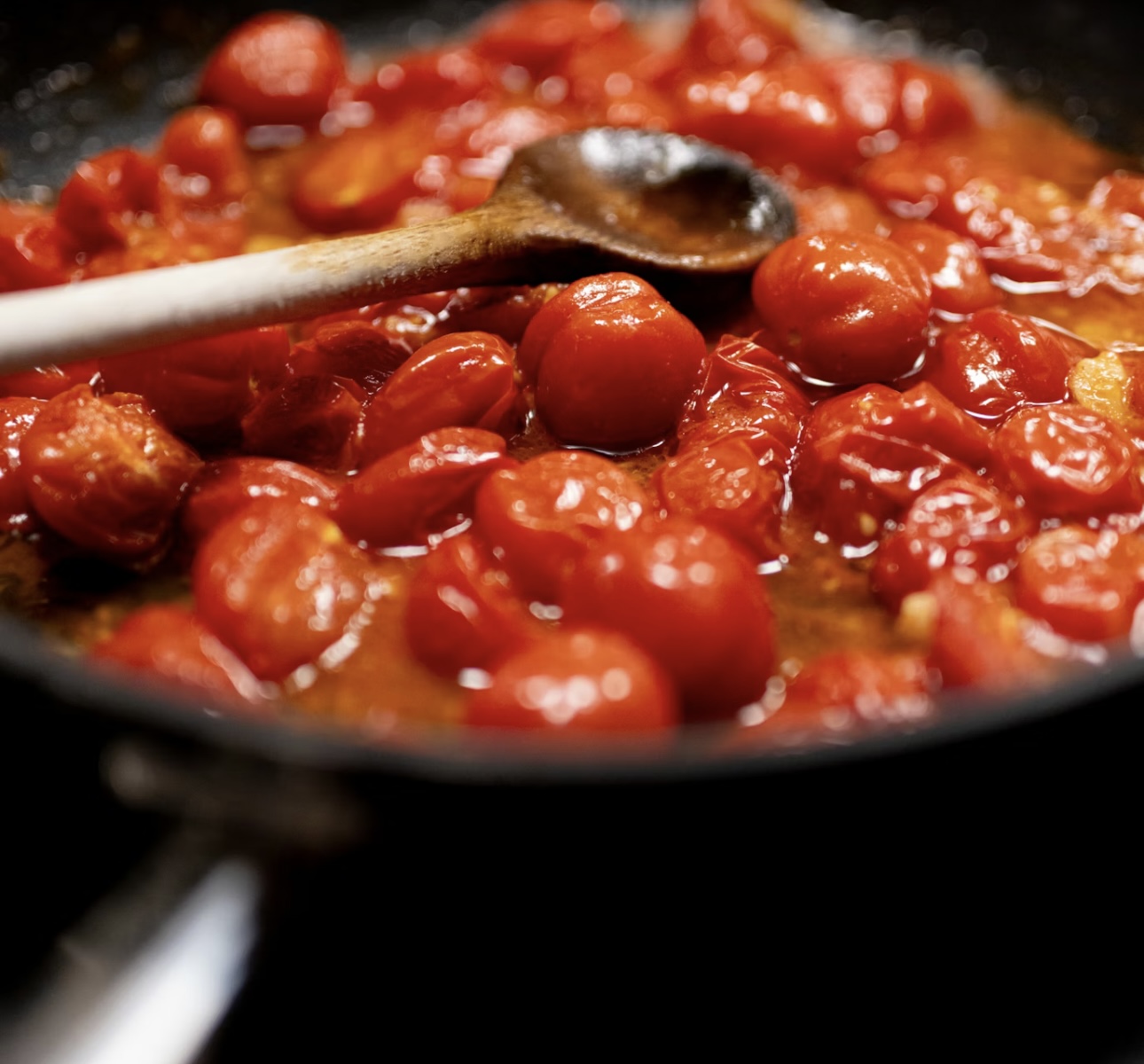 フライパンの中でミニトマトが煮崩れ、木べらで軽く混ぜられながらソース状になっていく様子。