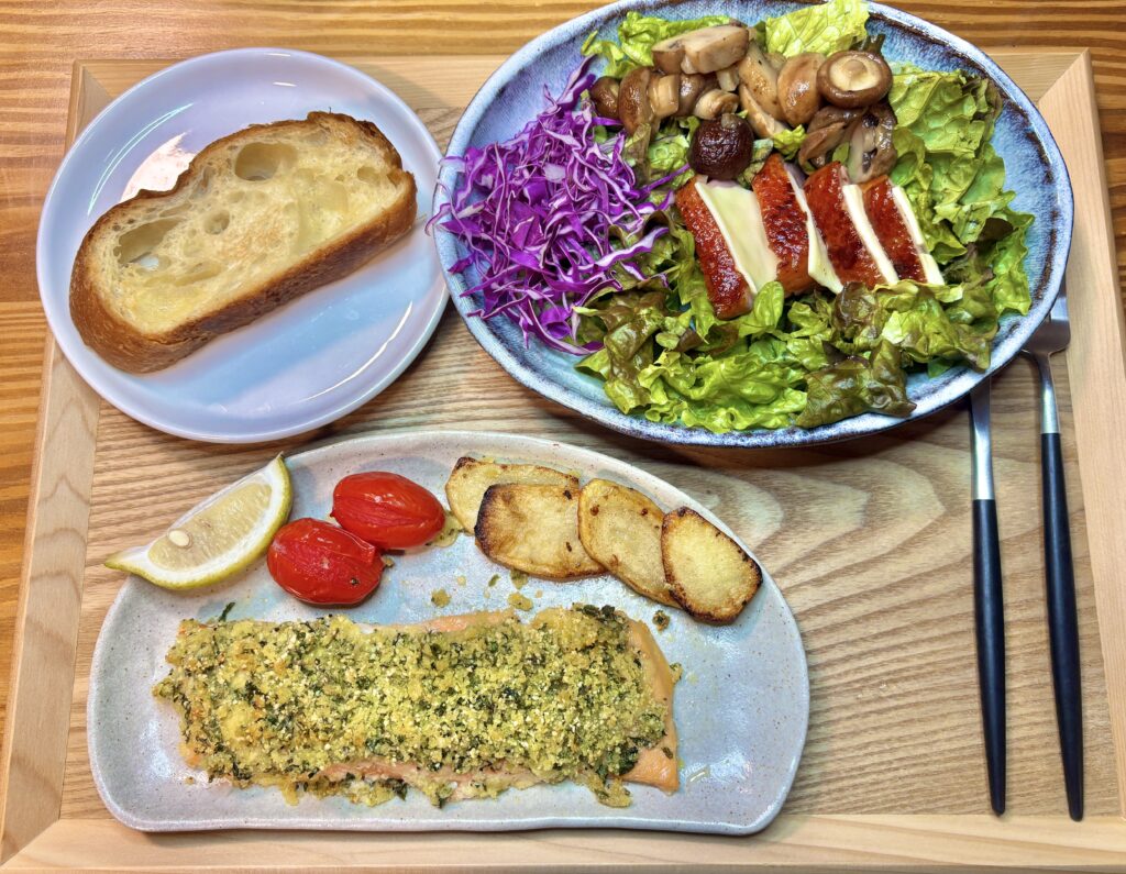 木製トレーに乗った豪華な夕食の献立全体写真。手前にはサーモンの香草パン粉焼きと付け合わせの野菜、奥には鴨のローストとチーズが乗った彩り豊かなサラダ、左にはパンが並んでいる。
