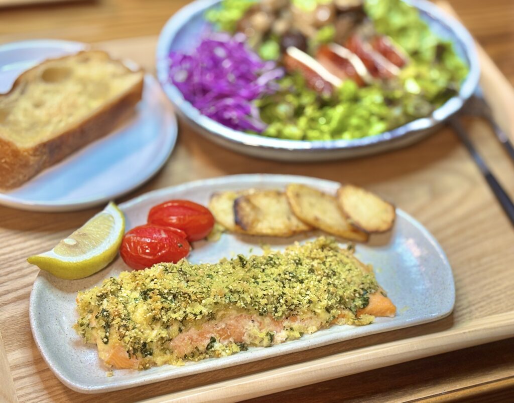 皿に盛られたサーモンの香草パン粉焼きの接写。こんがり焼けたパン粉の上にハーブが混ざっており、横にはローストしたプチトマト、ポテト、レモンが添えられている。
