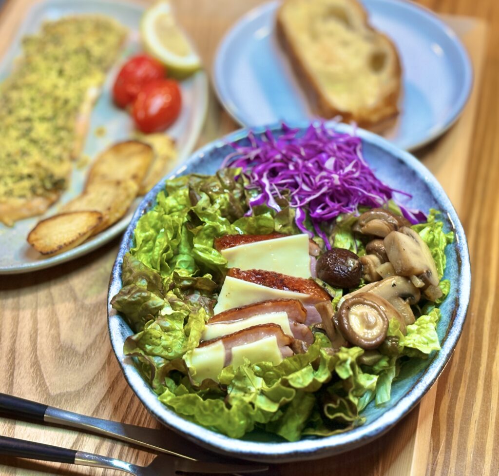 青い皿に盛られた彩り鮮やかなサラダの接写。レタスと紫キャベツの上に、スライスチーズを挟んだ鴨のローストと、ガーリックソテーされたキノコがたっぷりと乗せられている。
