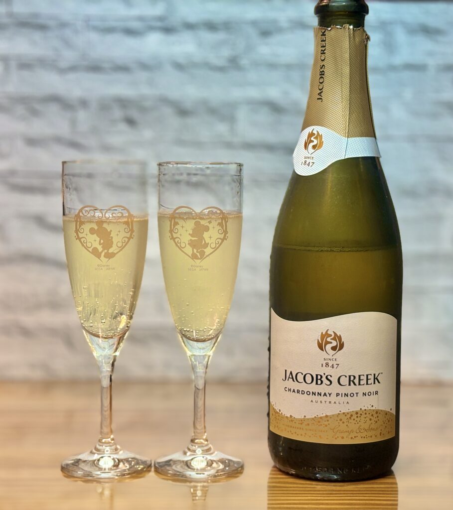 ジェイコブス・クリークのスパークリングワイン（シャルドネ・ピノ・ノワール）のボトルと、ワインが注がれた2つのシャンパングラス。グラスにはミッキーマウスとミニーマウスの金色の柄が施されている。
