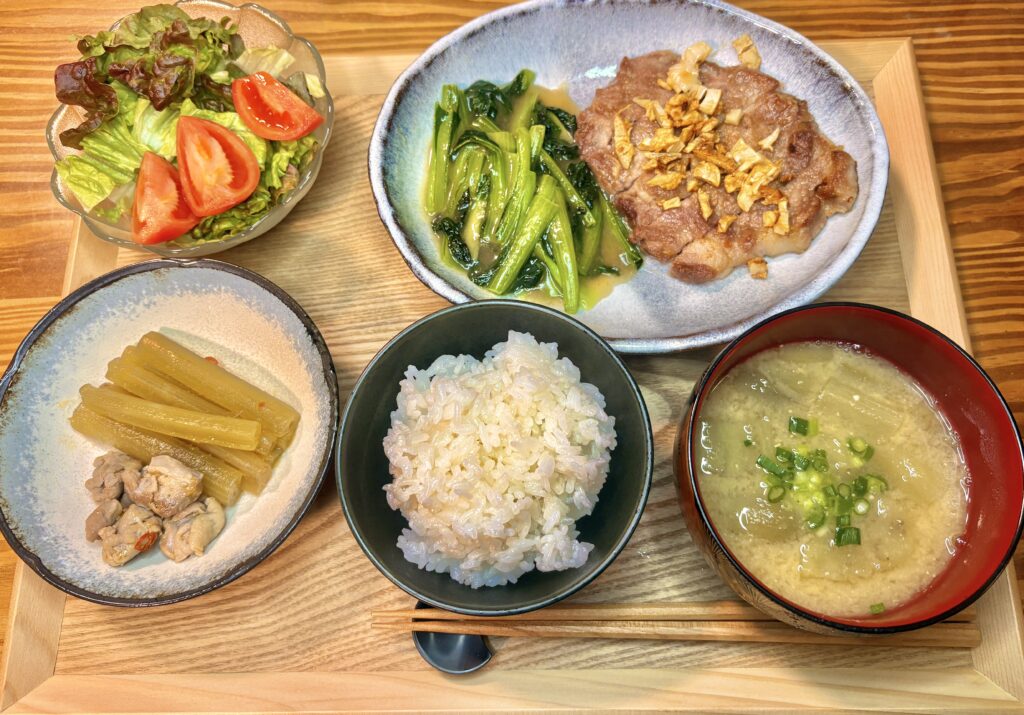 木製トレーの上に並んだ家庭の和定食。ご飯と焼きなす入りの味噌汁を中心に、ガーリックポークソテー、タアサイの中華炒め、ふきとかしわの煮物、レタスとトマトのサラダがバランスよく盛り付けられている
