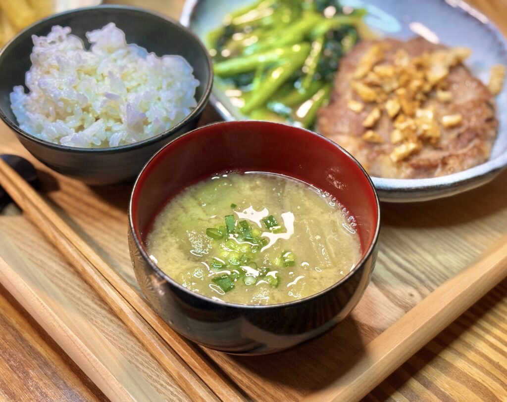 明るい木目調のテーブルに置かれた、ネギを散らした温かいお味噌汁。背景には彩り豊かなエディブルフラワーが添えられた前菜の皿。