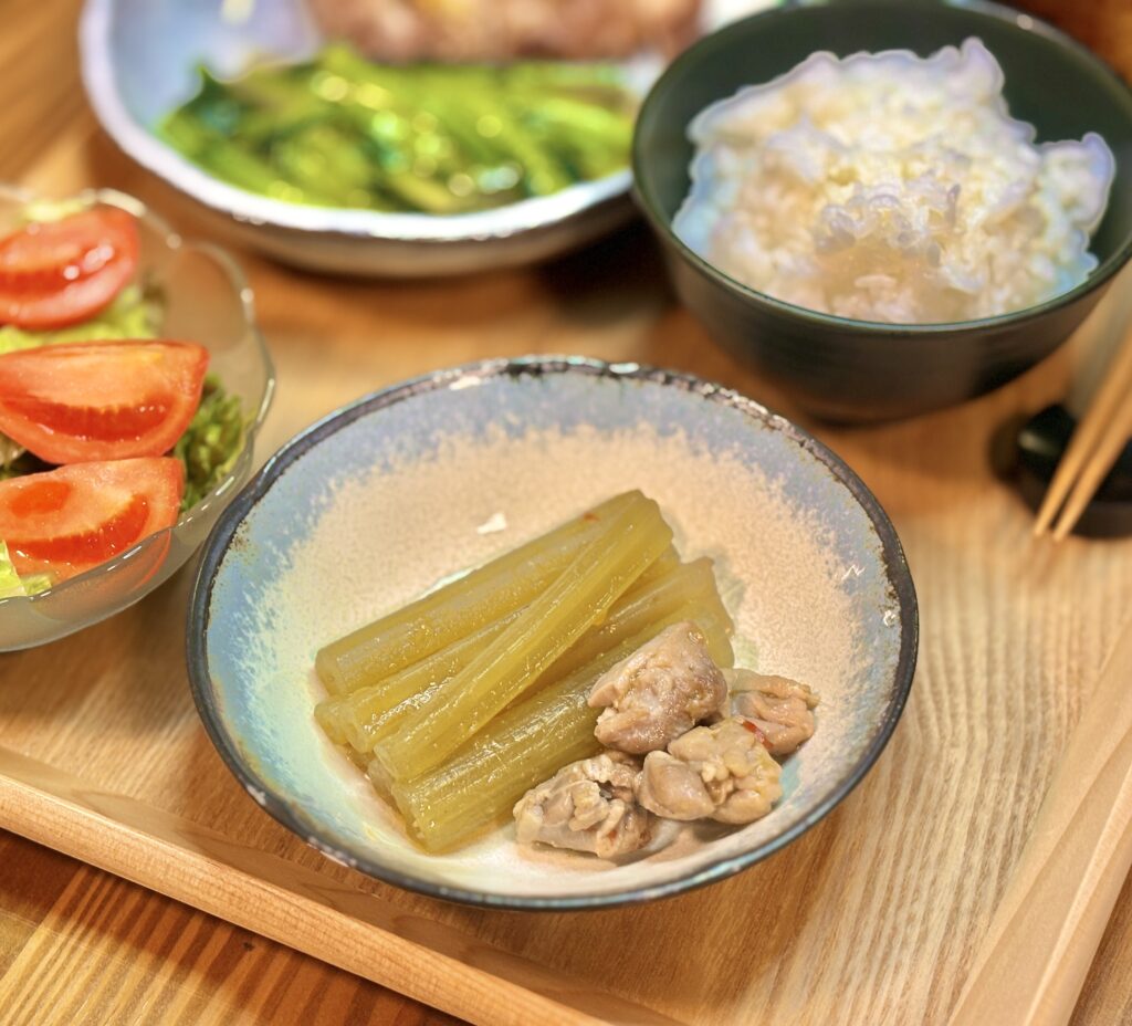 青い縁取りの小皿に盛り付けられた、出汁の染みたふきと豚肉の煮物。背景にお味噌汁やご飯が並ぶ食卓の風景。