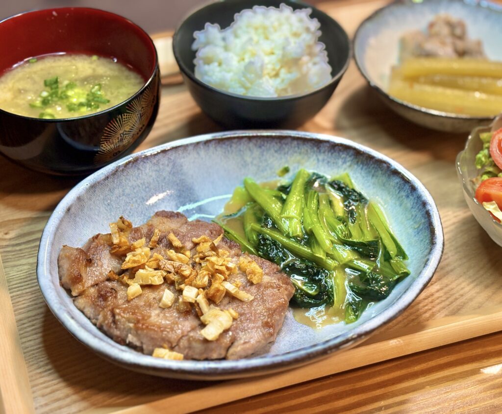 器に盛り付けられたガーリックポークソテーと、つやのあるタアサイの中華炒め。こんがり焼いた豚肉の上には刻みガーリックがのり、家庭的で食欲をそそる主菜の一皿