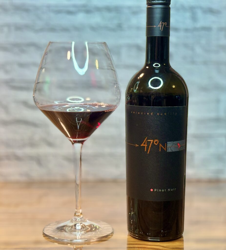 「47°N Selected quality Pinot Noir」のラベルが貼られた赤ワインのボトルと、ワインが注がれた大きなワイングラス。背景は明るいレンガ調の壁。