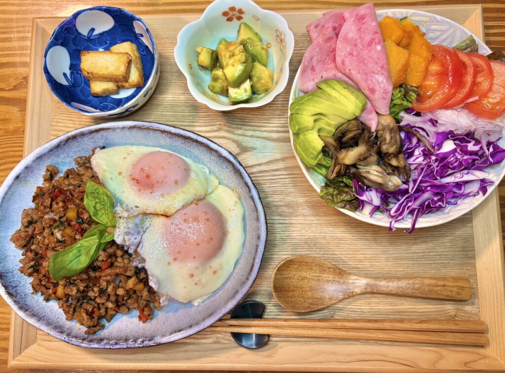 豚肉のガパオライスに目玉焼きを2枚のせた主菜と、厚揚げ、アボカドのわさび和え、アボカドとビアソーセージ、かぼちゃ、トマト、舞茸、紫キャベツ、レタスを盛り合わせたサラダが並ぶ木製トレーの食卓写真