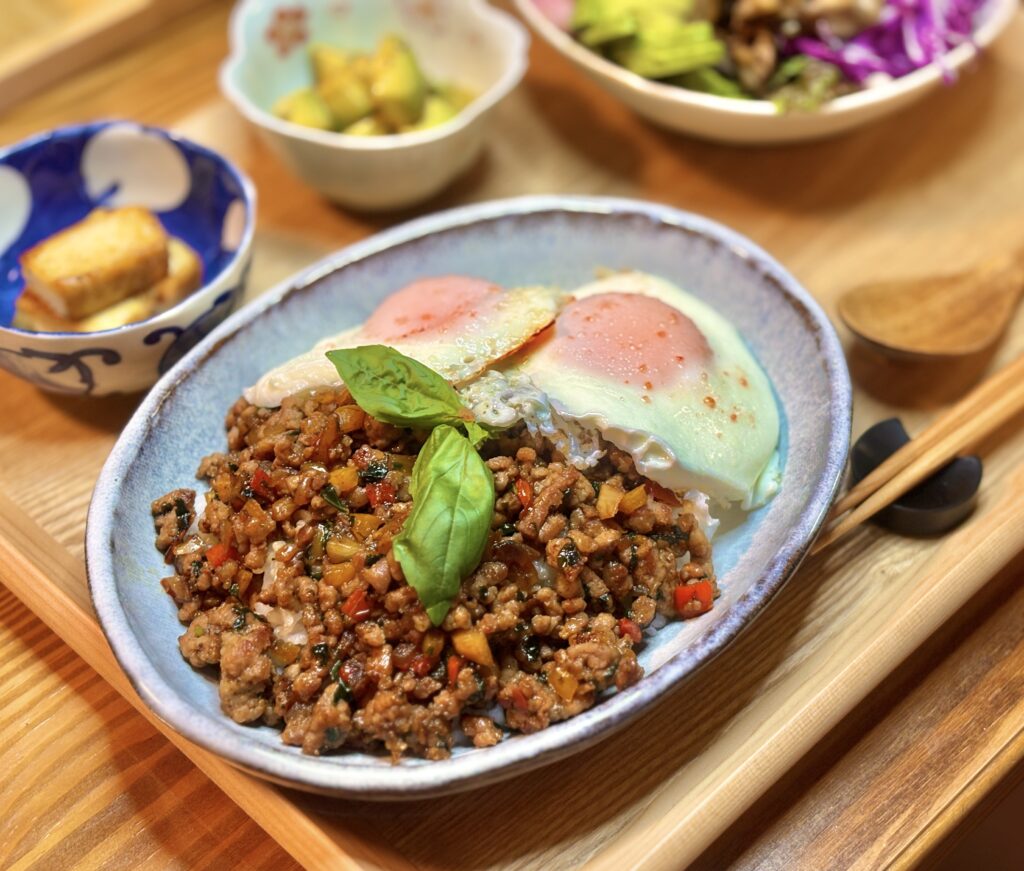 豚ひき肉のガパオライスに半熟の目玉焼きを2枚のせ、バジルを添えた主菜を木製トレーに盛り付けた写真。横に箸と小鉢の厚揚げが並ぶ温かみのある食卓風景