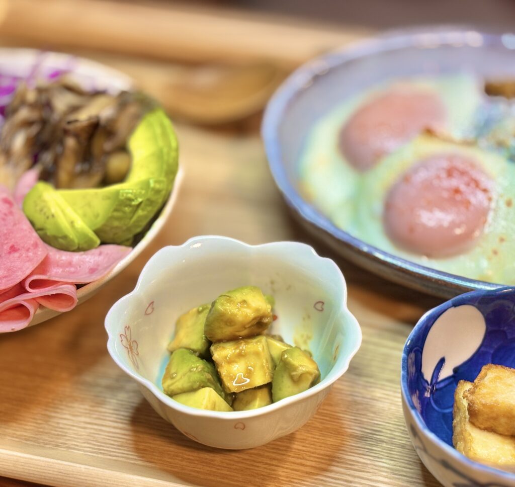 アボカドをわさびで和えた副菜を小鉢に盛り付け、奥に目玉焼きをのせた豚肉のガパオライス、横に焼き厚揚げが並ぶ木製トレーの晩ごはん写真