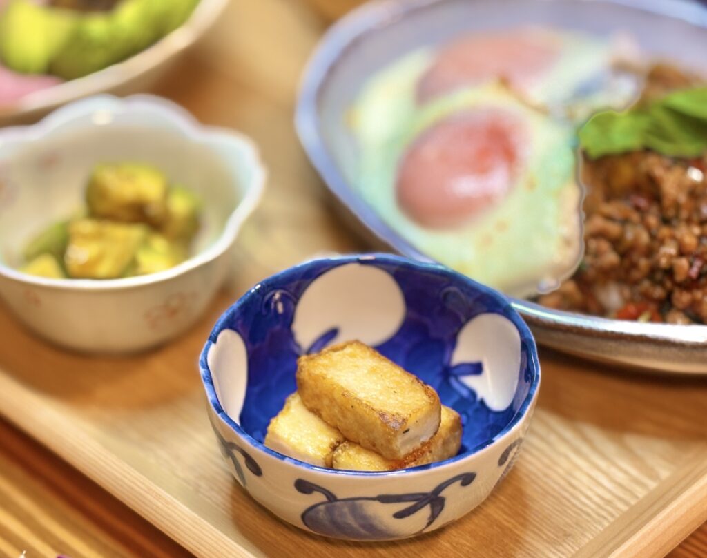 香ばしく焼いた厚揚げを小鉢に盛り付け、奥にアボカドのわさび和えと豚肉のガパオライス目玉焼きの主菜が並ぶ木製トレーの晩ごはん写真