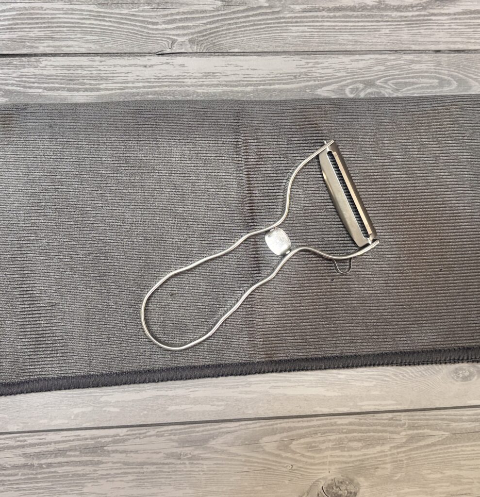 グレーのランチョンマットの上に置かれたステンレス製のピーラー。シンプルなデザインで、キッチン用品として清潔感がある。